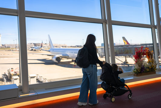 Une maman voyageant avec une poussette dans un aéroport, regardant les avions par la fenêtre avant un départ avec bébé.