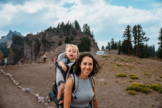 une maman porte son enfant sur son dos dans un porte bébé pendant une randonnée
